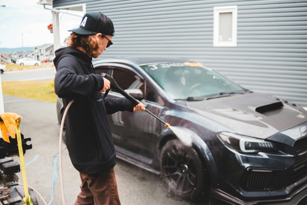 A man power washes a black sports car in a residential area, highlighting outdoor maintenance.